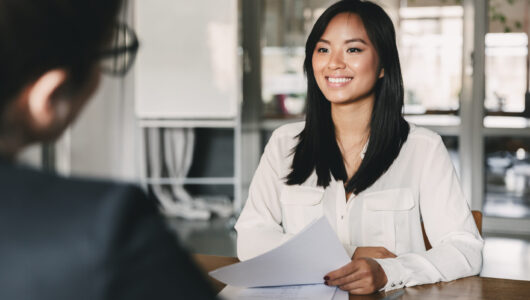 Portrait of joyful asian woman smiling and holding resume while sitting in front of businesswoman during corporate meeting or job interview - business, career and placement concept